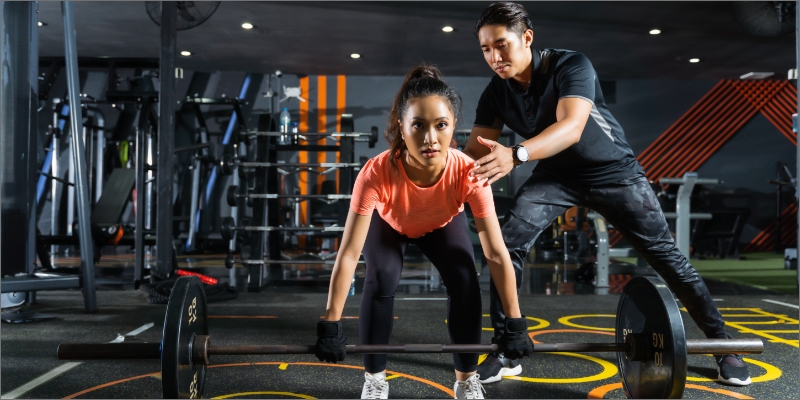 A young woman in deadlift posture in a gym room, and a gym personal trainer guides her through the exercise .