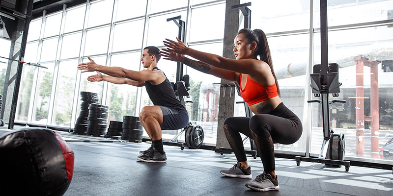 A physically fit woman and a guy is doing squats at the gym that is surrounded by exercise equipment.
